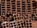 A close-up view of a stack of orange-brown rectangular hollow bricks, arranged in a pattern. The bricks have multiple square cavities running through them, designed for construction purposes. The lighting casts shadows within the cavities, highlighting the texture of the brick surface.