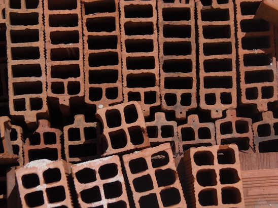 A close-up view of a stack of orange-brown rectangular hollow bricks, arranged in a pattern. The bricks have multiple square cavities running through them, designed for construction purposes. The lighting casts shadows within the cavities, highlighting the texture of the brick surface.