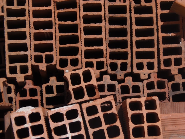A close-up view of a stack of orange-brown rectangular hollow bricks, arranged in a pattern. The bricks have multiple square cavities running through them, designed for construction purposes. The lighting casts shadows within the cavities, highlighting the texture of the brick surface.