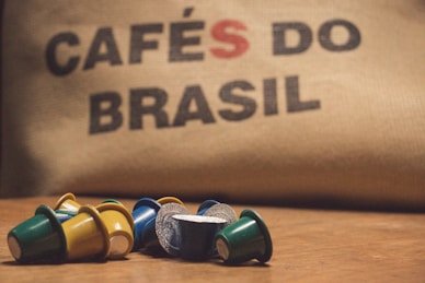 A collection of colorful coffee pods are scattered on a wooden surface in front of a burlap sack with the words 'CAFÉS DO BRASIL' printed on it.