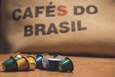 A collection of colorful coffee pods are scattered on a wooden surface in front of a burlap sack with the words 'CAFÉS DO BRASIL' printed on it.