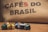A collection of colorful coffee pods are scattered on a wooden surface in front of a burlap sack with the words 'CAFÉS DO BRASIL' printed on it.