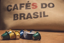 A collection of colorful coffee pods are scattered on a wooden surface in front of a burlap sack with the words 'CAFÉS DO BRASIL' printed on it.