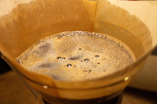 A close-up of freshly brewed coffee in a brown paper filter within a coffee dripper. The bubbling surface of the coffee features a foamy crema and dark coffee grounds. The texture and rich color indicate a freshly made pour-over coffee.