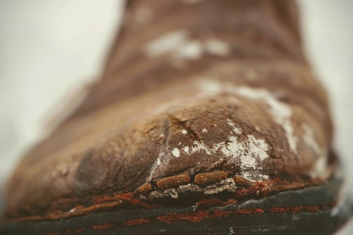 A close-up of a weathered and cracked piece of leather, possibly part of a shoe or boot, showing significant signs of wear with frayed edges and faded colors.