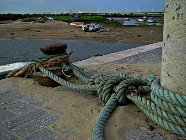 A mooring bollard with ropes tied around it is positioned on a sandy and paved surface near the edge of a body of water. In the background, a partially dried riverbed is visible with several small boats stranded on the sandy areas. The horizon showcases a strip of greenery and a few distant structures, suggesting a bridge or waterfront development.