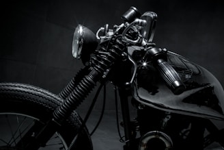 A close-up shot of a vintage motorcycle, emphasizing the details of the headlight, handlebar, and front tire. The image features dark and metallic tones, highlighting the intricate design and craftsmanship of the bike.