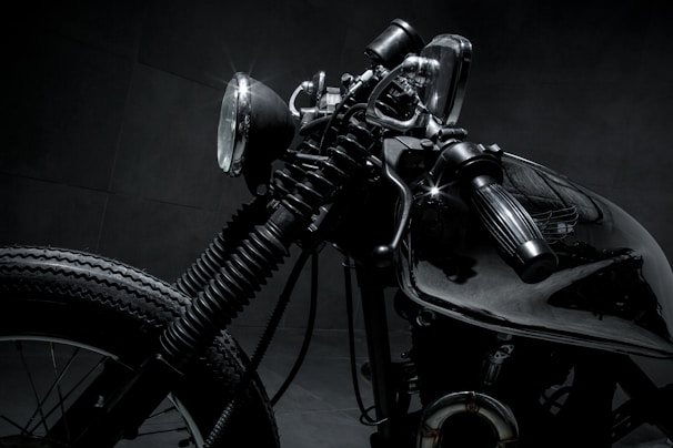 A close-up shot of a vintage motorcycle, emphasizing the details of the headlight, handlebar, and front tire. The image features dark and metallic tones, highlighting the intricate design and craftsmanship of the bike.