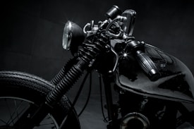 A close-up shot of a vintage motorcycle, emphasizing the details of the headlight, handlebar, and front tire. The image features dark and metallic tones, highlighting the intricate design and craftsmanship of the bike.