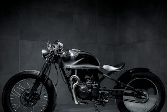 A sleek, custom-built motorcycle is positioned in a dimly lit room. The bike features a minimalist design with a shiny metallic finish and broad tires. The background is composed of dark tiles, lending a moody and industrial atmosphere to the scene.
