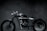 A sleek, custom-built motorcycle is positioned in a dimly lit room. The bike features a minimalist design with a shiny metallic finish and broad tires. The background is composed of dark tiles, lending a moody and industrial atmosphere to the scene.