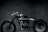 A sleek, custom-built motorcycle is positioned in a dimly lit room. The bike features a minimalist design with a shiny metallic finish and broad tires. The background is composed of dark tiles, lending a moody and industrial atmosphere to the scene.