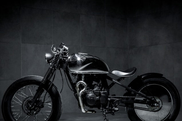A sleek, custom-built motorcycle is positioned in a dimly lit room. The bike features a minimalist design with a shiny metallic finish and broad tires. The background is composed of dark tiles, lending a moody and industrial atmosphere to the scene.