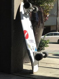A skateboard leaning against a wall in the shade with sunlight hitting the sidewalk. The skateboard has stickers including a red 'Almost' logo and 'Venice' text, alongside other graphic designs. A large potted plant is partially visible in the background, and a white car is parked on the street.