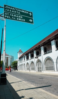 An urban street scene features a large historical white building with arched windows along the side. There is a clear, bright blue sky overhead. A street sign in the foreground points to different directions, with labels such as Johar/Tugumuda, Jakarta, St. Tawang, and Pel. TJ. Emas. A few vehicles, including a motorcycle, are on the road. There are also some modern buildings visible in the background.