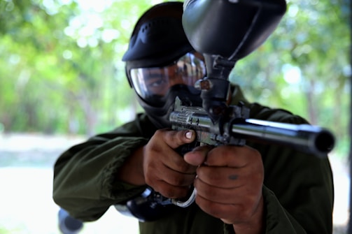 A person wearing protective goggles and a mask aims a paintball gun towards the camera. The setting appears to be outdoors with a blurred background of greenery. The individual is dressed in dark attire, emphasizing a focus on action or sport.