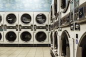 A laundromat with several rows of stacked industrial dryers. The machines have metallic finishes and display panels for user operation. The floor is tiled with a light beige color, and the upper wall features a sky pattern with clouds.