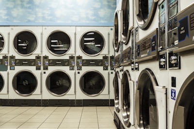 A laundromat with several rows of stacked industrial dryers. The machines have metallic finishes and display panels for user operation. The floor is tiled with a light beige color, and the upper wall features a sky pattern with clouds.