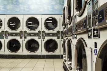 A laundromat with several rows of stacked industrial dryers. The machines have metallic finishes and display panels for user operation. The floor is tiled with a light beige color, and the upper wall features a sky pattern with clouds.