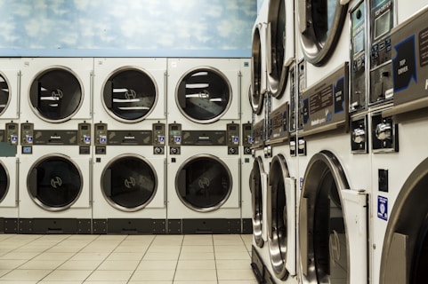 A laundromat with several rows of stacked industrial dryers. The machines have metallic finishes and display panels for user operation. The floor is tiled with a light beige color, and the upper wall features a sky pattern with clouds.
