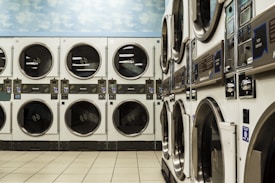 A laundromat with several rows of stacked industrial dryers. The machines have metallic finishes and display panels for user operation. The floor is tiled with a light beige color, and the upper wall features a sky pattern with clouds.