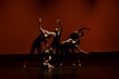 Five dancers are performing a contemporary dance routine on stage. They are dressed in black outfits, and their movements are dynamic and synchronized, creating a dramatic visual effect. The background is a warm orange-red color, enhancing the intensity of the scene.