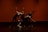 Five dancers are performing a contemporary dance routine on stage. They are dressed in black outfits, and their movements are dynamic and synchronized, creating a dramatic visual effect. The background is a warm orange-red color, enhancing the intensity of the scene.