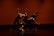 Five dancers are performing a contemporary dance routine on stage. They are dressed in black outfits, and their movements are dynamic and synchronized, creating a dramatic visual effect. The background is a warm orange-red color, enhancing the intensity of the scene.