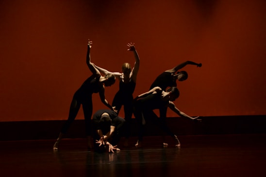 Five dancers are performing a contemporary dance routine on stage. They are dressed in black outfits, and their movements are dynamic and synchronized, creating a dramatic visual effect. The background is a warm orange-red color, enhancing the intensity of the scene.