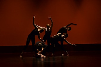 Five dancers are performing a contemporary dance routine on stage. They are dressed in black outfits, and their movements are dynamic and synchronized, creating a dramatic visual effect. The background is a warm orange-red color, enhancing the intensity of the scene.