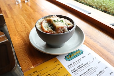 A wooden table in a cafe holds a gray bowl and plate, filled with a soup topped with melted cheese, herbs, and slices of bread. Next to the bowl is a menu from the Levure Bakery and Patisserie featuring various items and prices.