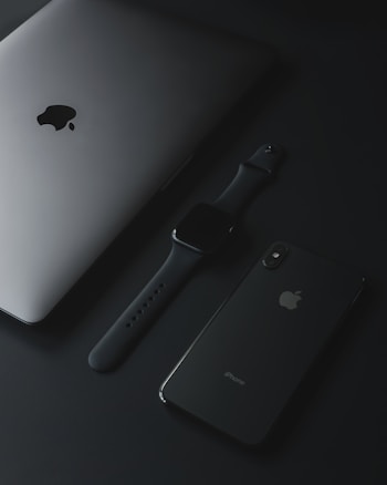 A sleek arrangement of Apple products resting on a dark surface, including a silver laptop, a smartwatch with a black band, and a modern smartphone. The devices are positioned artistically and exhibit a minimalist and sophisticated design aesthetic.