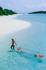 A woman wearing a hat and a swimsuit walking in clear shallow water, pulling a transparent kayak with two orange floatation devices. The beach is sandy and white, lined with lush greenery, and there is a distant view of more greenery and the horizon.