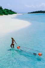 A woman wearing a hat and a swimsuit walking in clear shallow water, pulling a transparent kayak with two orange floatation devices. The beach is sandy and white, lined with lush greenery, and there is a distant view of more greenery and the horizon.