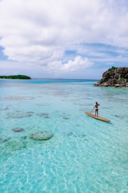 A person is standing on a paddleboard, gliding over crystal-clear turquoise water. The surface of the water is dotted with rocks and coral visible beneath. To the right, there is a rocky outcrop with green foliage atop, and in the distance, a small, lush green island is visible under a partly cloudy sky.