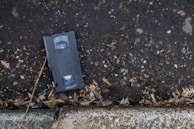 A VHS cassette sits on an asphalt surface next to a concrete curb. The ground is covered with small pebbles and scattered debris, including some dry twigs and leaves.