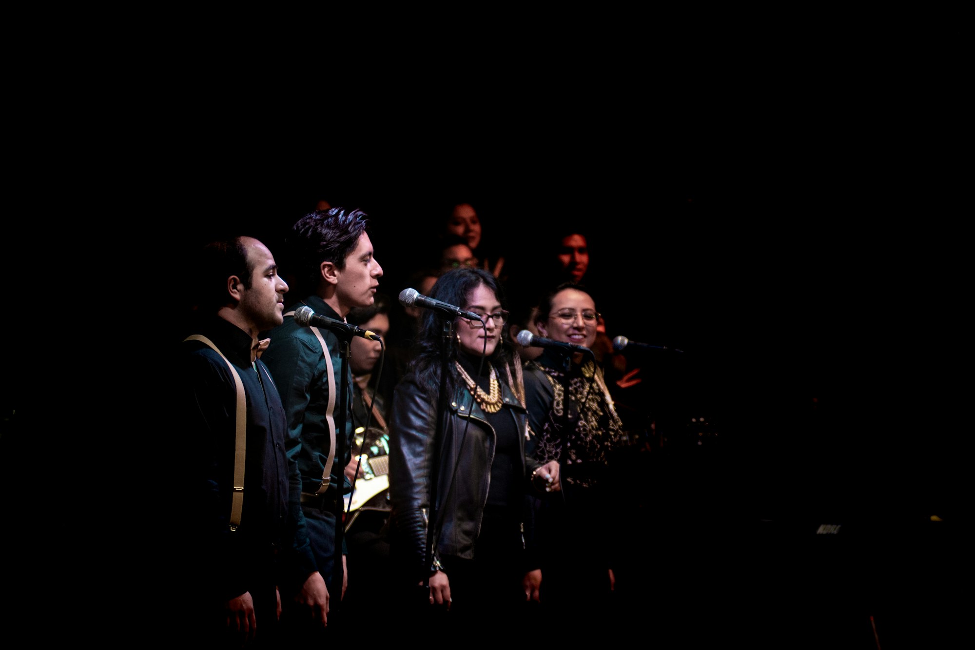 A small adult chorus sings into microphones at a concert.