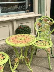 A decorative outdoor table and chair set featuring intricate rose patterns is positioned outside a shop with a sign that reads 'art and antiques'. The furniture is painted in a light green color with hints of pink on the roses. A small potted plant with orange flowers is placed on the ledge behind the table.