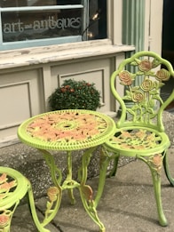 A decorative outdoor table and chair set featuring intricate rose patterns is positioned outside a shop with a sign that reads 'art and antiques'. The furniture is painted in a light green color with hints of pink on the roses. A small potted plant with orange flowers is placed on the ledge behind the table.