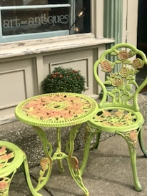 A decorative outdoor table and chair set featuring intricate rose patterns is positioned outside a shop with a sign that reads 'art and antiques'. The furniture is painted in a light green color with hints of pink on the roses. A small potted plant with orange flowers is placed on the ledge behind the table.