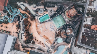 Aerial view of a water park featuring multiple slides in turquoise and dark colors laid out over a sandy terrain. Several pools with varying shapes and sizes are visible, surrounded by pathways and landscaping elements. The park is bordered by buildings and roads.