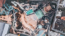 Aerial view of a water park featuring multiple slides in turquoise and dark colors laid out over a sandy terrain. Several pools with varying shapes and sizes are visible, surrounded by pathways and landscaping elements. The park is bordered by buildings and roads.