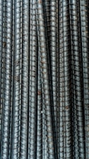 Close-up view of multiple parallel steel rebar rods with a textured, ribbed surface. The bars have a weathered and industrial appearance, indicating they are used in construction settings.