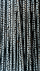 Close-up view of multiple parallel steel rebar rods with a textured, ribbed surface. The bars have a weathered and industrial appearance, indicating they are used in construction settings.