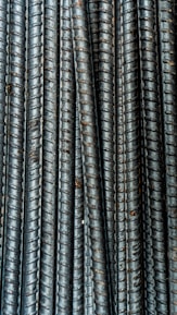Close-up view of multiple parallel steel rebar rods with a textured, ribbed surface. The bars have a weathered and industrial appearance, indicating they are used in construction settings.