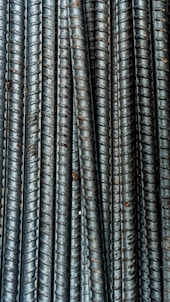 Close-up view of multiple parallel steel rebar rods with a textured, ribbed surface. The bars have a weathered and industrial appearance, indicating they are used in construction settings.
