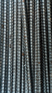 Close-up view of multiple parallel steel rebar rods with a textured, ribbed surface. The bars have a weathered and industrial appearance, indicating they are used in construction settings.
