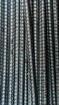 Close-up view of multiple parallel steel rebar rods with a textured, ribbed surface. The bars have a weathered and industrial appearance, indicating they are used in construction settings.