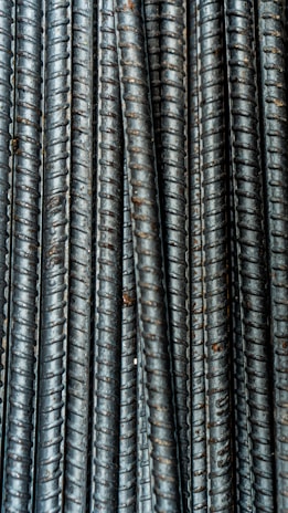 Close-up view of multiple parallel steel rebar rods with a textured, ribbed surface. The bars have a weathered and industrial appearance, indicating they are used in construction settings.