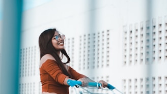 A young woman with long dark hair and glasses is smiling while riding a bicycle. She is dressed in an off-shoulder orange top and appears to be enjoying herself. There is a modern building with a lattice design in the background.